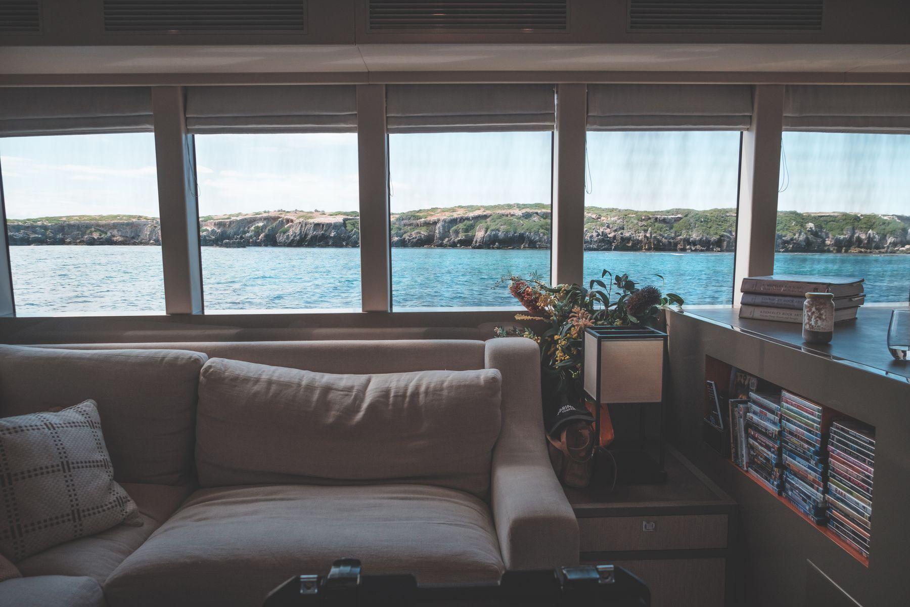 A yacht salon with a grey sofa beneath panoramic windows onto turquoise water